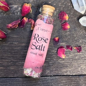 May include: A small glass bottle with a cork stopper filled with pink rose salt and dried rose petals. The bottle has a pink label that reads "of Forest and Fire Rose Salt ritual salt".