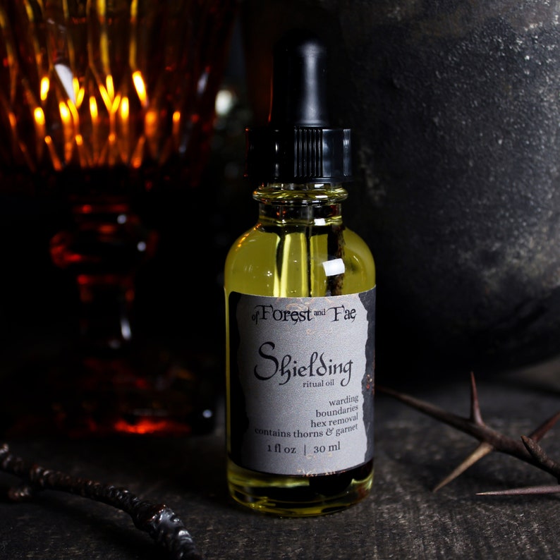 May include: A small glass bottle with a black dropper top and a black and white label that reads "of Forest and Fae Shielding ritual oil warding boundaries hex removal contains thorns & garnet 1 fl oz | 30 ml".