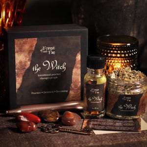 May include: A black box labeled Forest of and Fae the Witch with a 0.5 fl oz bottle of anointing oil, a jar of dried herbs, a metallic taper candle, three polished red and brown stones, and a copper-colored skeleton key talisman.