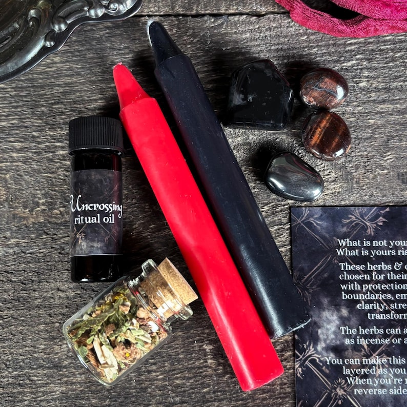 May include: A black and red candle set, a small bottle of oil labeled "Uncrossing ritual oil", a small jar of dried herbs, and several black and brown stones. The text on the card reads "What is not yours rise, What is yours rise, These herbs & crystals are chosen for their protective properties with protection for boundaries, empowerment, clarity, strength, transformation. The herbs can be used as incense or a bath. You can make this layered as you wish. When you're ready, reverse side."