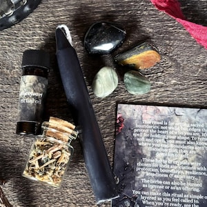 K&ouml;nnte beinhalten: Eine Sammlung von Ritualgegenst&auml;nden, darunter eine schwarze Kerze, eine kleine Flasche "Wermut-Ritual&ouml;l", ein Glas mit Kr&auml;utern und mehrere Kristalle. Eine Karte mit Text ist ebenfalls enthalten. Die Gegenst&auml;nde sind auf einer Holzoberfl&auml;che angeordnet.