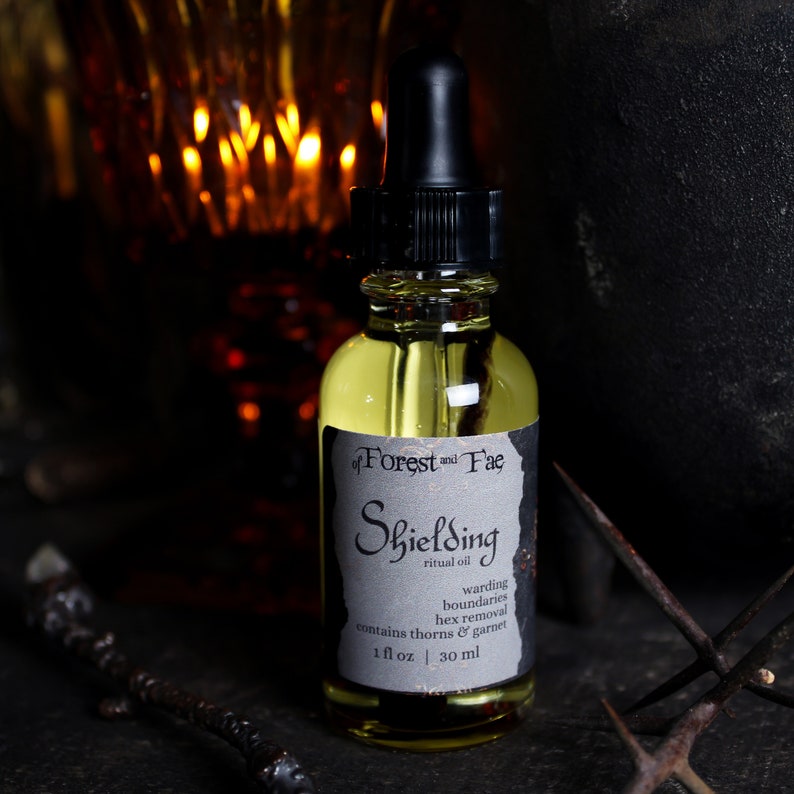 May include: A small amber glass bottle with a black dropper top and a white label that reads "of Forest and Fae Shielding ritual oil warding boundaries hex removal contains thorns & garnet 1 fl oz | 30 ml".