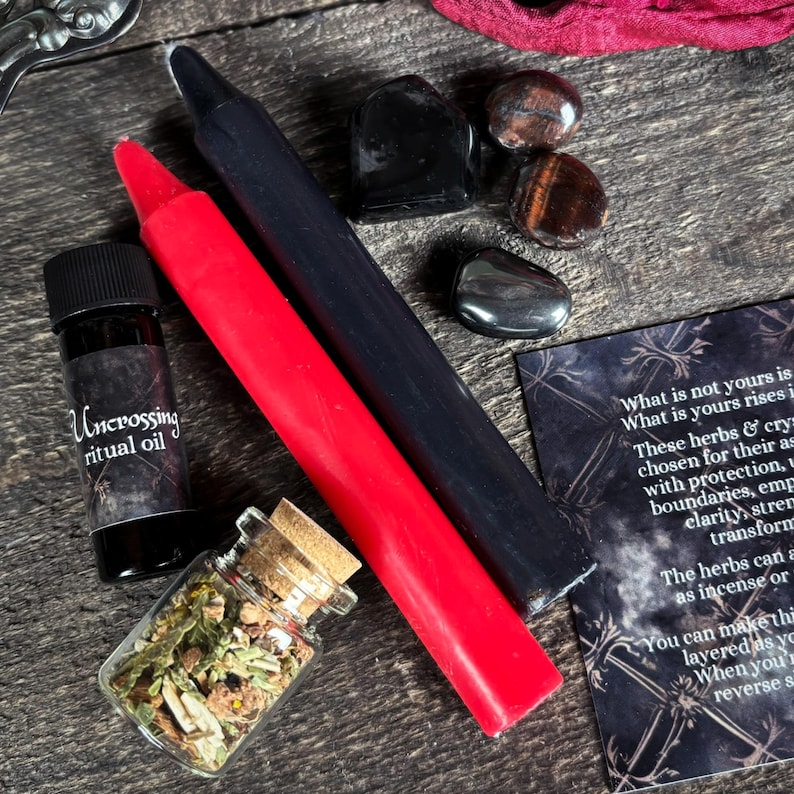 May include: A red and black candle set, a small bottle of oil labeled "Uncrossing ritual oil", a small jar of dried herbs, and four crystals: black obsidian, tiger's eye, and two hematite stones. The crystals are arranged on a wooden surface. A card with text about the herbs and crystals is visible in the background.