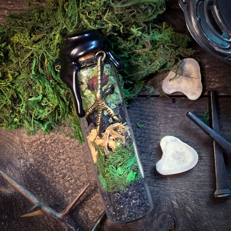 May include: A glass bottle with a black wax seal top filled with green moss, black sand, and a gold metal bird foot charm. The bottle is sitting on a wooden surface with moss and bone-like objects.