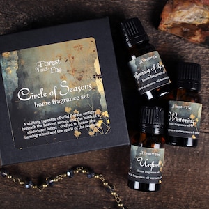 May include: A home fragrance set with a black box featuring the text "Circle of Seasons" and "Forest and Fae." Three small amber glass bottles of home fragrance oil are visible, labeled "Unfurl," "Wintering," and "Beginning of the." A decorative necklace is in the foreground.