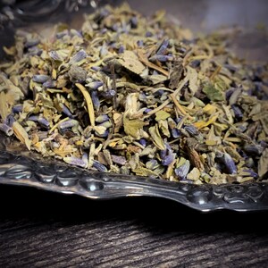 May include: A close-up of a silver bowl filled with dried herbs and lavender flowers. The herbs are a mix of green and brown, and the lavender flowers are a light purple.