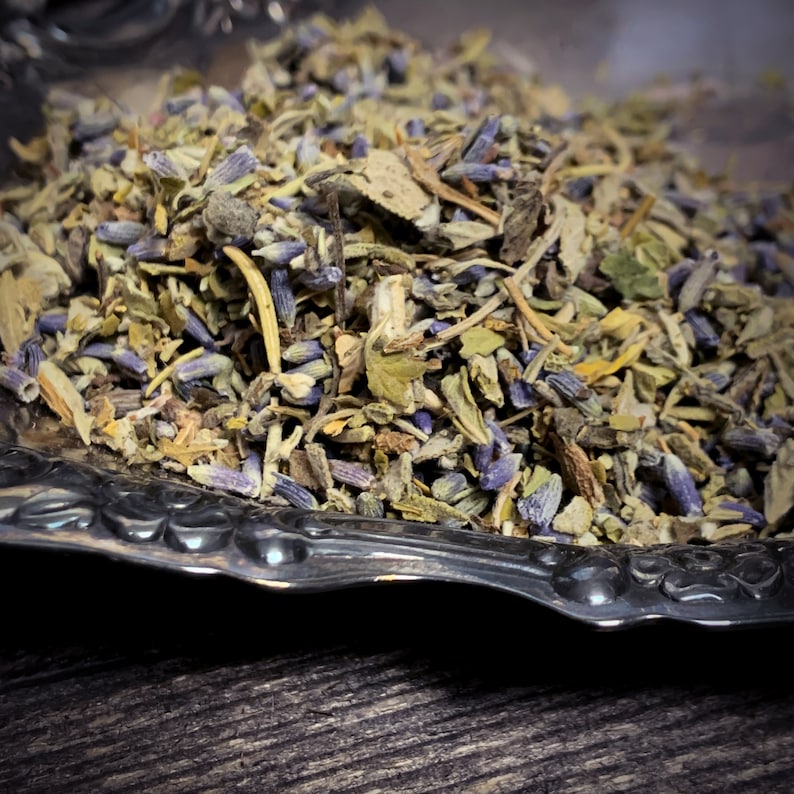 May include: A close-up of a silver bowl filled with dried herbs and lavender flowers. The herbs are a mix of green and brown, and the lavender flowers are a light purple.