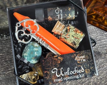 May include: A black box with a silver key, an orange candle, a small bottle of oil, a small jar of herbs, and a green stone. The box has a label that says "Unlocked road opening kit" and "Forest of the Fae".