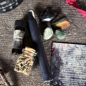 K&ouml;nnte beinhalten: Eine Sammlung spiritueller Gegenst&auml;nde, darunter eine schwarze Kerze, Kristalle, eine kleine Flasche Ritual&ouml;l und ein Glasgef&auml;&szlig; mit Kr&auml;utern. Die Gegenst&auml;nde sind auf einer Holzoberfl&auml;che angeordnet, mit einer Karte mit Text &uuml;ber Rituale.