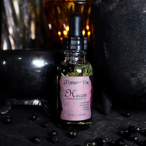 May include: A small glass bottle of Hecate sacred ritual oil with a dropper top. The bottle is filled with a clear liquid and has a pink and black label with the text "of Forest and Fae Hecate sacred ritual oil crossroads dark moon shadow hedge 1 fl oz | 30 ml".