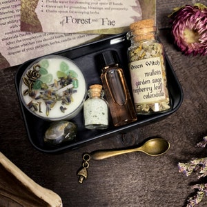 May include: A black metal box with a charm spoon, a small candle, a bottle of salt, a bottle of oil, and a jar of dried herbs. The box is labeled "Green Witch" and lists the herbs included: mullein, garden sage, raspberry leaf, and calendula.