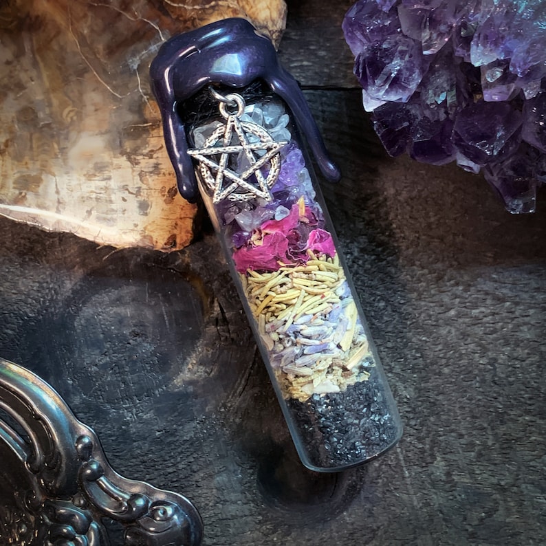 Puede incluir: Un vial de vidrio lleno de varios cristales, hierbas y flores secas. El vial est&aacute; coronado con un sello de cera negra y un amuleto de pentagrama plateado.