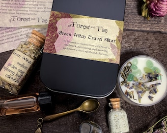 May include: A black metal box with the text "of Forest and Fae Green Witch Travel Altar" on the front. The box contains a variety of items, including a small jar of herbs, a bottle of oil, a spoon, a small jar of salt, a candle, and a small charm.