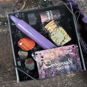 May include: A Crossroads spell kit with a purple candle, a small bottle of herbs, a red and orange agate stone, and a card that says "Crossroads spell kit Forest of and Fae".