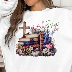 May include: White sweatshirt featuring a watercolor design with a wooden cross, stacked books, flowers, and mushrooms. The text "In Jesus' Name" is written in script. The design has a religious theme.