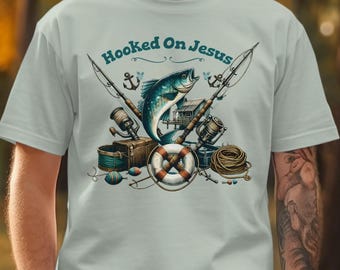 Könnte beinhalten: Hellblaues T-Shirt mit einem Angelmotiv. Das Design umfasst einen großen blauen Fisch, Angelruten, eine Rettungsboje und den Text "Hooked On Jesus" in Türkis. Ein lässiges Kleidungsstück.