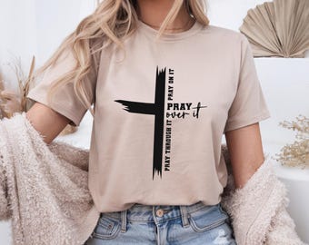 May include: A beige t-shirt featuring a black cross design with the words "Pray Over It" and "Pray Through It" printed on it. The shirt is made of a soft material and has short sleeves.