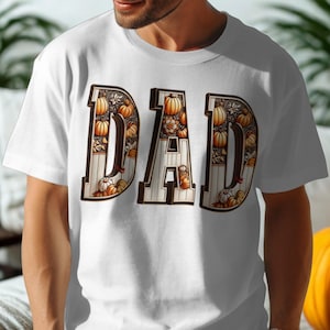 May include: White t-shirt featuring the word "DAD" in large, decorative letters. The letters are filled with a pumpkin and floral design, with a brown border. The shirt is made of a soft material.