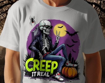 May include: White t-shirt with a skeleton wearing sunglasses and a black jacket, seated before a purple moon and bats. The shirt has the text "CREEP IT REAL" in green and yellow, with a pumpkin and spider.