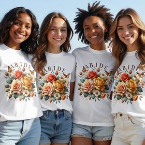 Op de afbeelding: Vier mensen dragen witte t-shirts met het woord "ABIDE" boven een bloemenontwerp van rozen en vlinders. De rozen zijn in tinten geel, oranje en rood. De mensen lachen en dragen spijkerbroekshorts.