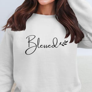 May include: White sweatshirt featuring the word "Blessed" in black cursive script, with a small leaf detail. The sweatshirt is a simple, casual style.