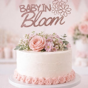 Könnte beinhalten: Eine weiße Torte, verziert mit rosa Rosen aus Zuckerguss und belegt mit frischen Blumen und einem glitzernden "Baby in Bloom" Tortenaufsatz. Die Torte steht auf einem weißen Kuchenständer, ideal für eine Babyparty.