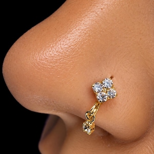 May include: A gold nose ring featuring a cluster of four clear, round-cut stones arranged in a square shape, with a single dangling charm consisting of a smaller, matching clear stone attached by a small gold jump ring to the main setting.