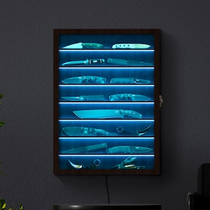 Puede incluir: Una vitrina rectangular de pared con marco de madera oscura y siete estantes horizontales. Cada estante está iluminado por una tira de luz LED azul brillante, exhibiendo varios cuchillos dispuestos en filas tras un panel frontal de cristal.
