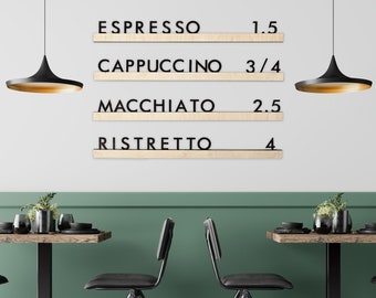 May include: A wall-mounted coffee menu sign featuring four light wood shelves with black 3D lettering. The text reads ESPRESSO 1.5, CAPPUCCINO 3/4, MACCHIATO 2.5, and RISTRETTO 4. The sign is displayed above two small tables with black chairs.