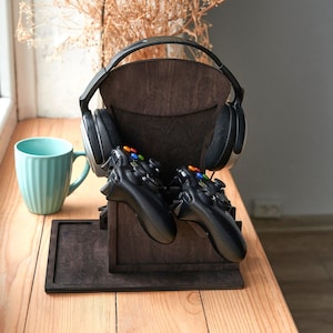 Peut inclure: Un support de casque de jeu en bois brun foncé avec deux manettes de jeu noires et un casque noir. Une tasse bleu clair est à gauche. Le support est sur une surface en bois.