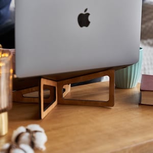 K&ouml;nnte beinhalten: Ein Laptopst&auml;nder aus Holz mit hellbrauner Oberfl&auml;che und geometrischem Design. Der St&auml;nder h&auml;lt einen silbernen Laptop mit einem Apple-Logo. Der Laptop steht auf einem Holztisch mit einer Glaslampe und Watteb&auml;uschen.