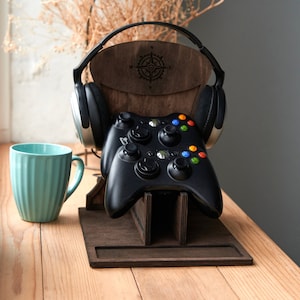 Pode incluir: Um suporte de madeira com um design de bússola segura dois controladores de videogame pretos e um par de fones de ouvido pretos. O suporte está em uma superfície de madeira clara.