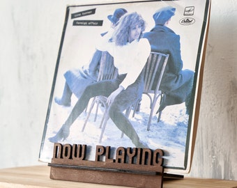 May include: A dark brown wooden display stand with the words NOW PLAYING cut out in a bold font. The stand holds a square vinyl record sleeve featuring a blue-toned photograph of a person sitting on a chair in a snowy outdoor setting.