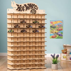 May include: A light-coloured wooden display rack featuring multiple horizontal shelves with individual compartments for small toy cars. The top of the unit is shaped with the Hot Wheels logo. Several small toy cars are placed on the upper shelves.