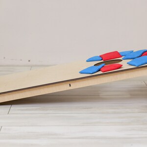 Puede incluir: Un tablero de juego cornhole de madera clara con un orificio circular en la parte superior, inclinado sobre un soporte. Varios sacos de tela rectangulares peque&ntilde;os en colores rojo y azul s&oacute;lidos est&aacute;n esparcidos por la superficie.