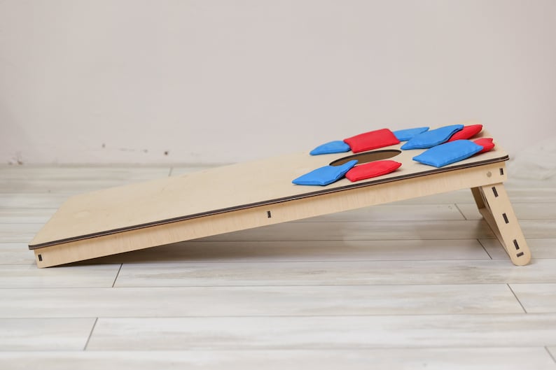 Puede incluir: Un tablero de juego cornhole de madera clara con un orificio circular en la parte superior, inclinado sobre un soporte. Varios sacos de tela rectangulares peque&ntilde;os en colores rojo y azul s&oacute;lidos est&aacute;n esparcidos por la superficie.