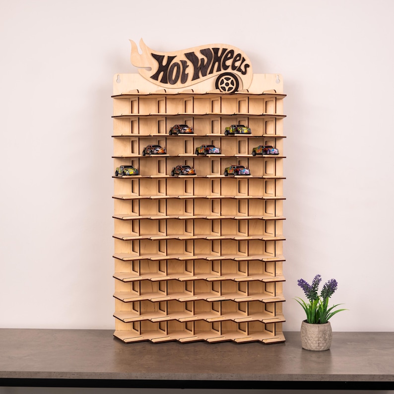 May include: A light-coloured wooden wall-mounted display rack featuring multiple horizontal shelves designed for small toy cars. The top of the unit displays the Hot Wheels logo cut out in wood. Several small toy cars are placed on the upper shelves.
