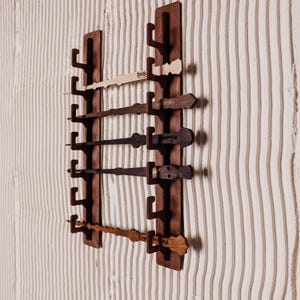 Può includere: Un porta bacchette da parete in legno con sei slot. Il supporto è marrone scuro e ha una finitura in legno naturale. Le bacchette sono in legno e hanno colori e design diversi. Le bacchette sono etichettate con nomi come "Reddie", "Expelliarmus" e "Petrificus Totalus".