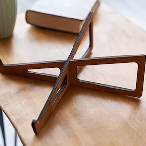 K&ouml;nnte beinhalten: Ein Laptop-St&auml;nder aus Holz mit dunkelbrauner Oberfl&auml;che. Der St&auml;nder besteht aus zwei Holzst&uuml;cken, die in der Mitte miteinander verbunden sind und ein X bilden. Der St&auml;nder ist so konzipiert, dass er einen Laptop auf eine ergonomischere H&ouml;he anhebt.