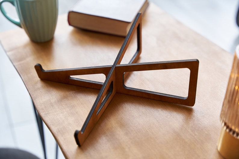 K&ouml;nnte beinhalten: Ein Laptop-St&auml;nder aus Holz mit dunkelbrauner Oberfl&auml;che. Der St&auml;nder besteht aus zwei Holzst&uuml;cken, die in der Mitte miteinander verbunden sind und ein X bilden. Der St&auml;nder ist so konzipiert, dass er einen Laptop auf eine ergonomischere H&ouml;he anhebt.
