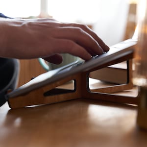 K&ouml;nnte beinhalten: Ein Laptopst&auml;nder aus Holz mit einem schwarzen Laptop darauf. Der St&auml;nder ist abgewinkelt, um ergonomische Unterst&uuml;tzung f&uuml;r den Laptop zu bieten.