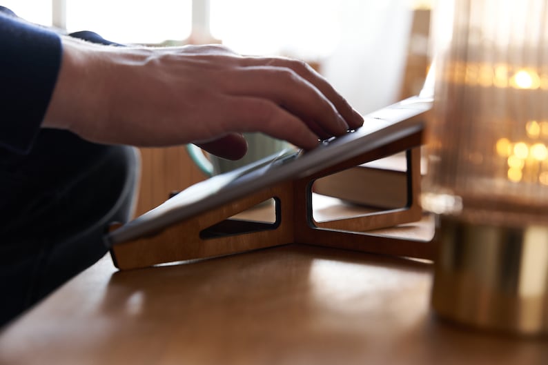K&ouml;nnte beinhalten: Ein Laptopst&auml;nder aus Holz mit einem schwarzen Laptop darauf. Der St&auml;nder ist abgewinkelt, um ergonomische Unterst&uuml;tzung f&uuml;r den Laptop zu bieten.