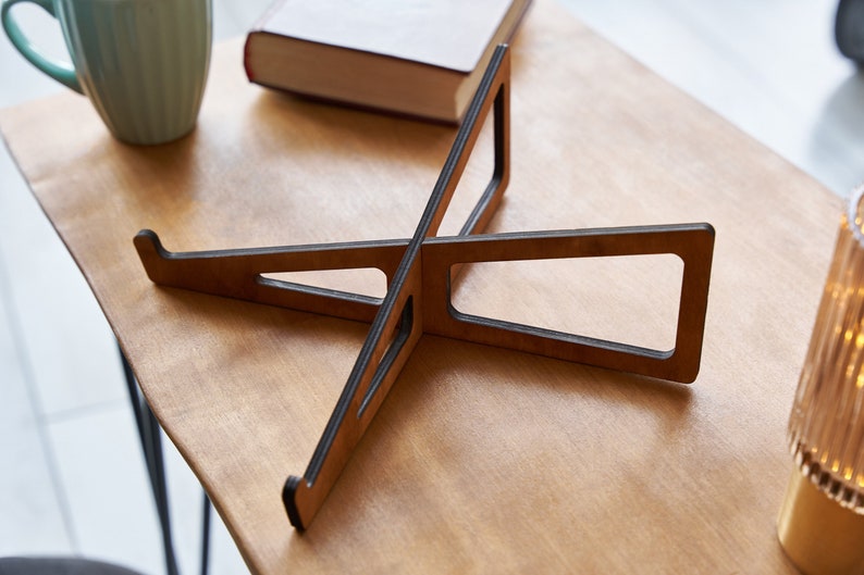 K&ouml;nnte beinhalten: Ein Laptop-St&auml;nder aus Holz mit dunkelbrauner Oberfl&auml;che. Der St&auml;nder besteht aus zwei Holzst&uuml;cken, die in der Mitte miteinander verbunden sind, um eine X-Form zu bilden. Der St&auml;nder ist so konzipiert, dass er einen Laptop auf eine ergonomischere Betrachtungsh&ouml;he anhebt.