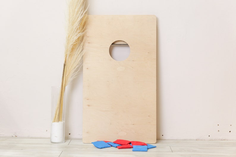 Puede incluir: Un tablero de cornhole rectangular de madera clara que presenta un orificio circular cerca del centro superior. En la base del tablero hay varias bolsas de frijoles cuadradas peque&ntilde;as de tela en colores rojo y azul, dispuestas en un mont&oacute;n.
