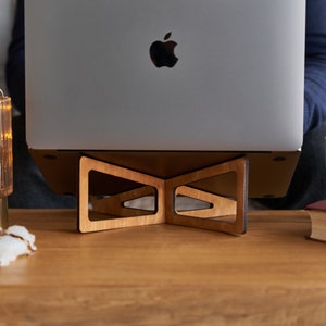 K&ouml;nnte beinhalten: Ein Laptopst&auml;nder aus Holz mit einem dreieckigen Design, der einen geschlossenen Laptop mit einem Apple-Logo auf dem Deckel tr&auml;gt. Der St&auml;nder steht auf einem Holztisch mit einer Lampe, Watteb&auml;uschen und einem Buch.