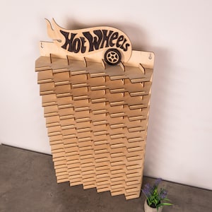 May include: A light-coloured wooden wall-mounted display rack featuring a Hot Wheels logo at the top with a dark wood accent. The rack has multiple horizontal shelves with notched slots designed for organising and showcasing small toy vehicles.