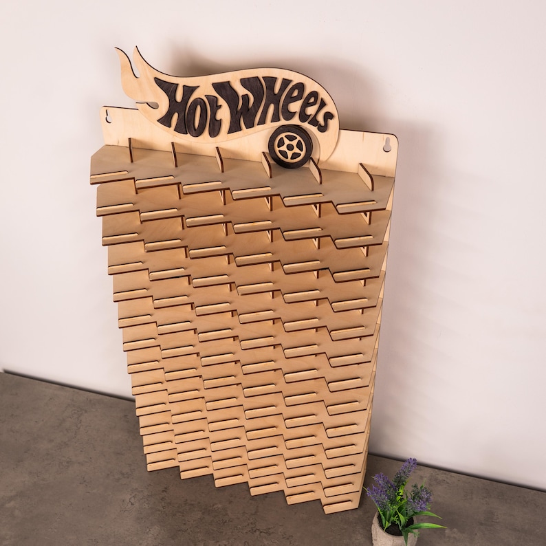 May include: A light-coloured wooden wall-mounted display rack featuring a Hot Wheels logo at the top with a dark wood accent. The rack has multiple horizontal shelves with notched slots designed for organising and showcasing small toy vehicles.