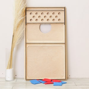 Puede incluir: Un tablero de cornhole rectangular de madera clara natural con bordes negros. El tablero incluye una secci&oacute;n superior con doce orificios circulares y un gran agujero central. Varias bolsas de tela peque&ntilde;as de color rojo y azul se sit&uacute;an debajo.