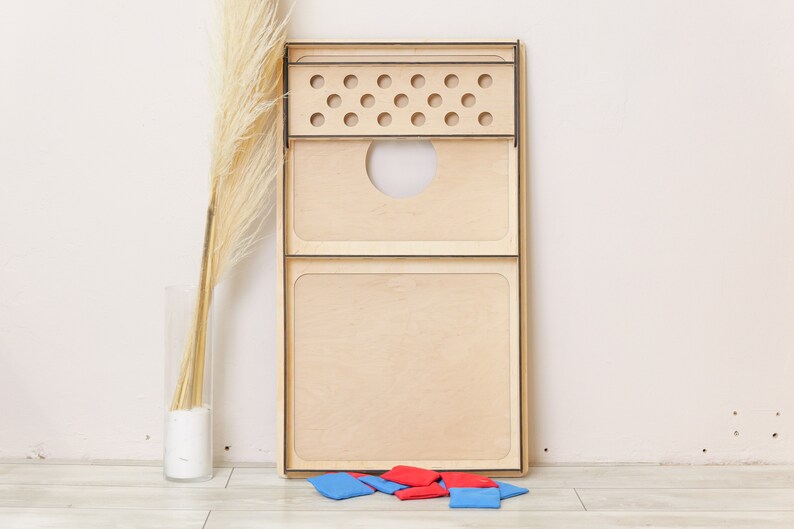 Puede incluir: Un tablero de cornhole rectangular de madera clara natural con bordes negros. El tablero incluye una secci&oacute;n superior con doce orificios circulares y un gran agujero central. Varias bolsas de tela peque&ntilde;as de color rojo y azul se sit&uacute;an debajo.
