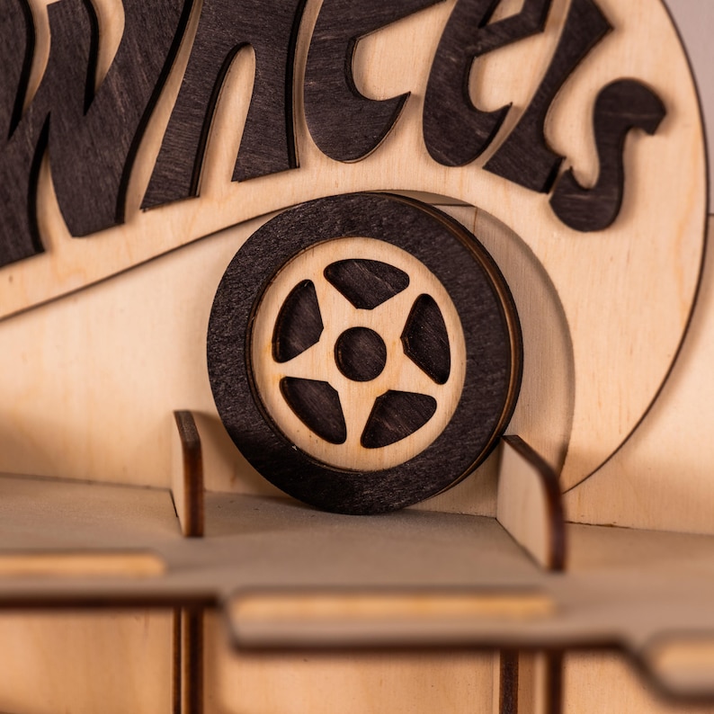 May include: A laser-cut wooden display shelf featuring a dark stained wheel icon and the partial text "WHEELS" in dark wood lettering. The structure is made of light-coloured plywood with multiple horizontal tiers and vertical dividers for organisation.